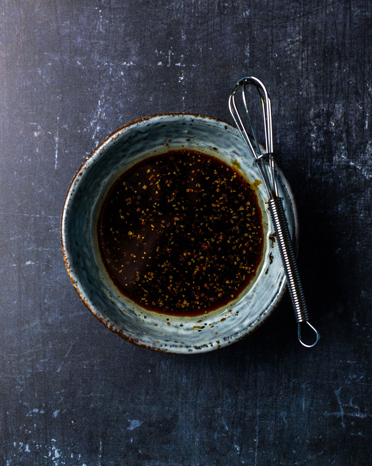 A small blue bowl is sitting on a dark tabletop. The bowl is filled with a dark salad dressing and there's a mini whisk on the side.