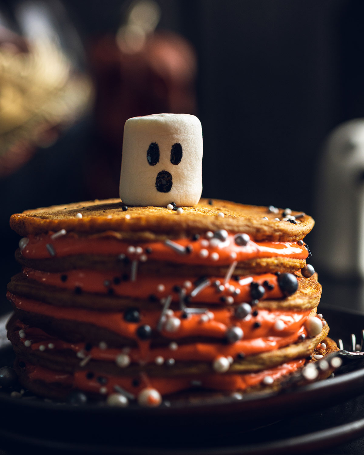 A stack of vegan pumpkin spice pancakes on a black plate topped with a ghost shaped marshmallow