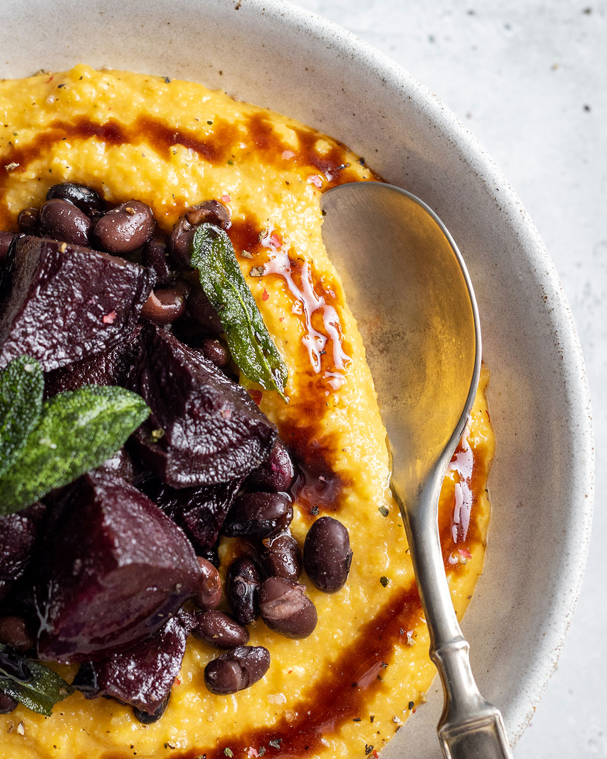 A white bowl filled with creamy pumpkin polenta topped with roasted beetroot and black beans in a balsamic sauce. On top of the bowl there are crunchy fresh sage leaves and a balsamic drizzle.