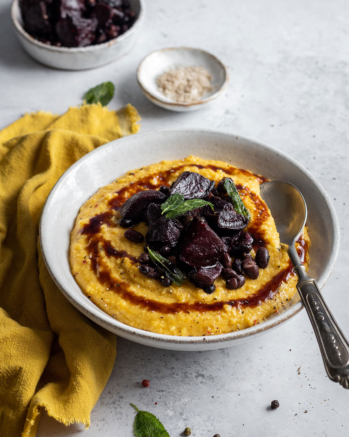 A full shot of a white ball and a white background next to a yellow cloth filled with creamy pumpkin polenta in a yellow colour. The pumpkin polenta is topped with roasted beetroot black beans and a balsamic drizzle.