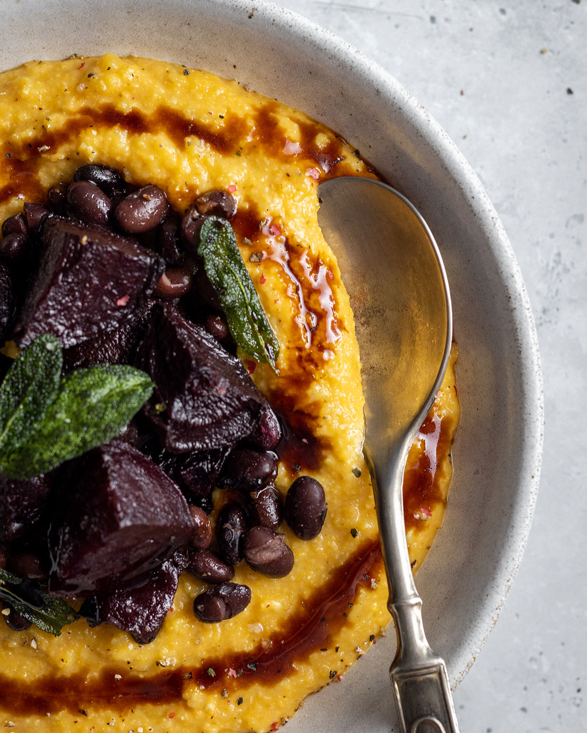 A white bowl with a spoon filled with yellow creamy pumpkin polenta made to a vegan recipe. The pumpkin polenta is topped with roasted beetroot black beans and a balsamic drizzle as well as crispy sage leaves.