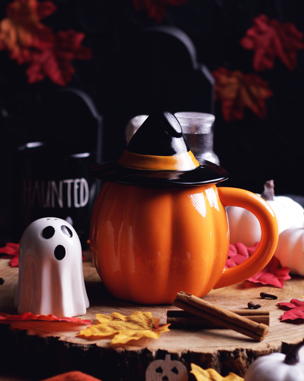 a decorated pumpkin mug with a black hat topper, a small ceramic ghost next to it with black gravestones in the background