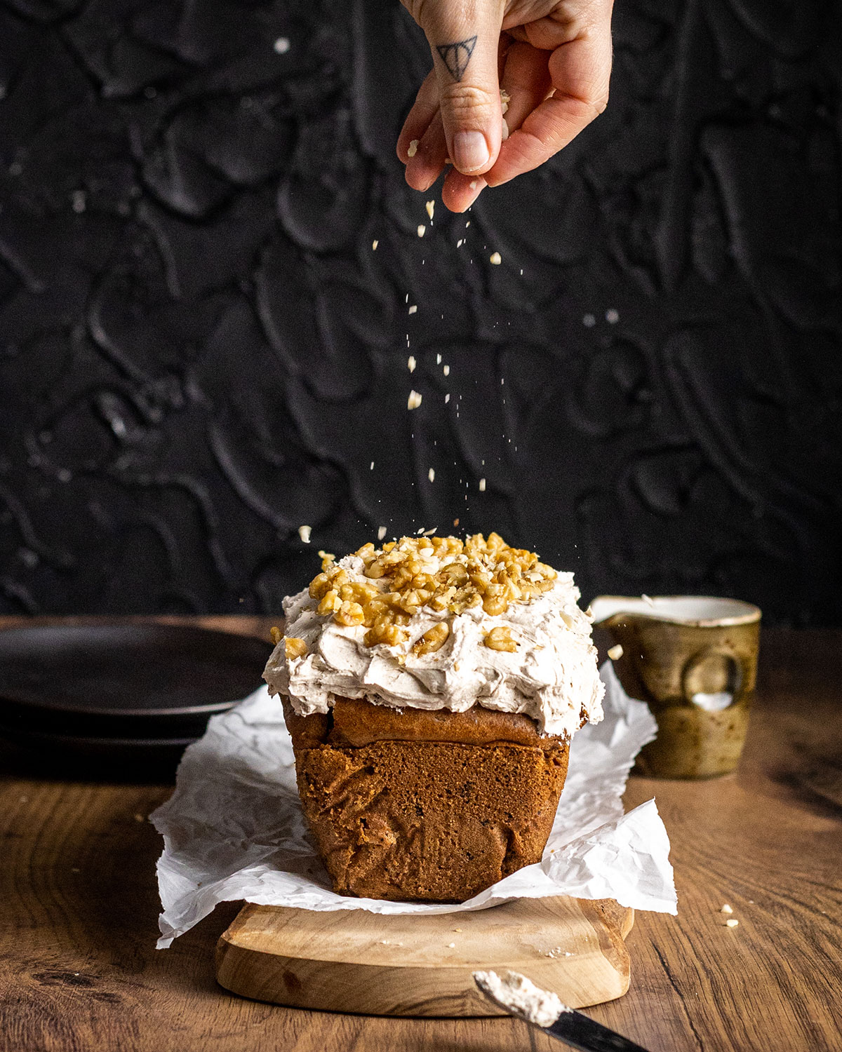 A hand sprinkling nuts on top of a whole loaf of coffee and walnut loaf cake in front of a black backdrop.
