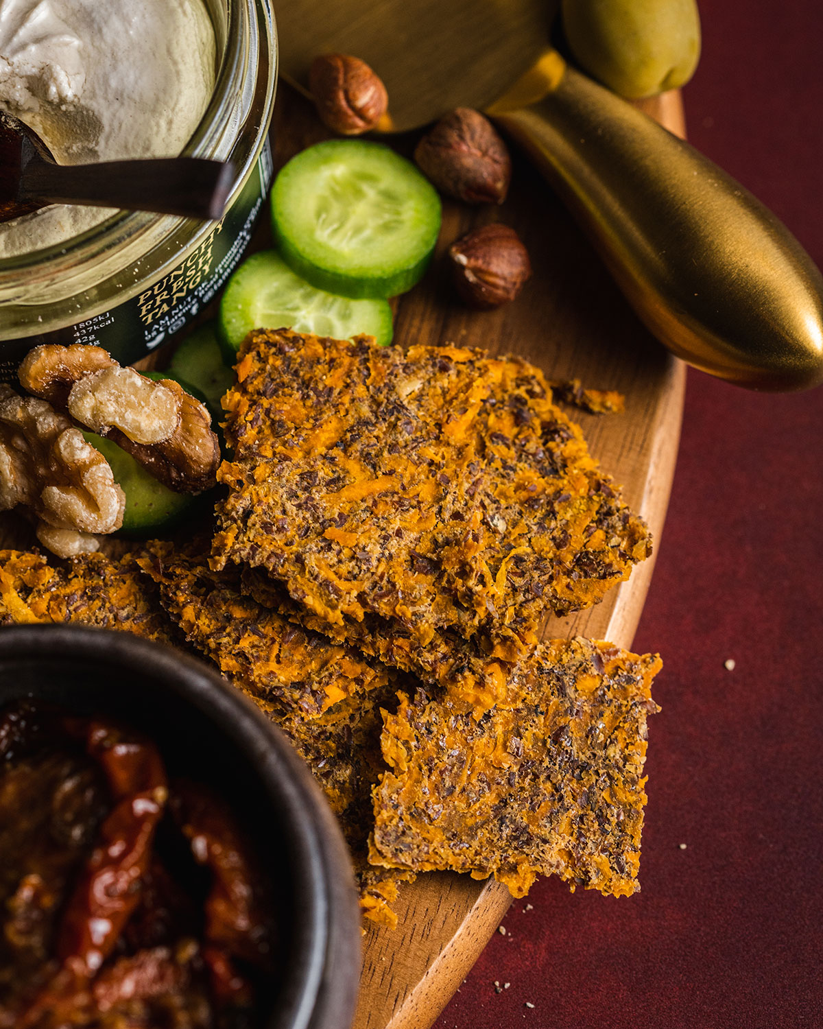 A close up photo of a vegan cheeseboard corner with homemade sweet potato crackers piled up on the edge, a cheese knife, sundried tomatoes, nuts and cucumbers places alongside.
