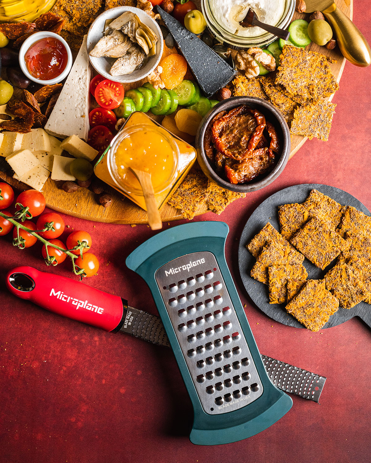 The edge of a large vegan cheeseboard with a plate of sweet potato crackers and both the Microplane Bowl Grater and Microplace Zester Grater placed next to it. 