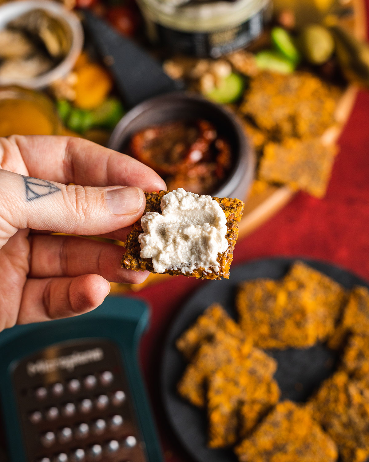 a hand holding a single sweet potato cracker that is topped with vegan ricotta. In the background you can see a blurry cheeseboard,