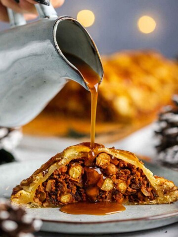 Vegan Wellington stuffed with vegetables and beans, a slice sitting on a light plate with the full vegan Wellington in the background and a hand drizzling vegan gravy over the vegan Wellington.