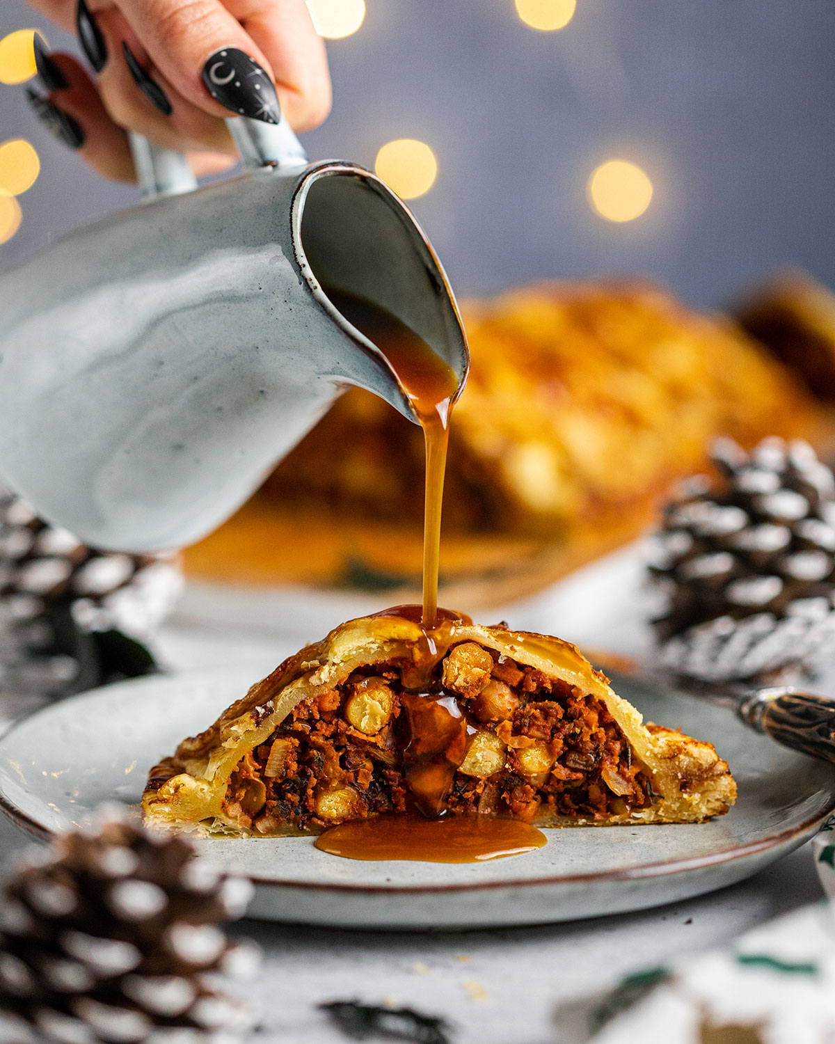 Vegan Wellington stuffed with vegetables and beans, a slice sitting on a light plate with the full vegan Wellington in the background and a hand drizzling vegan gravy over the vegan Wellington.