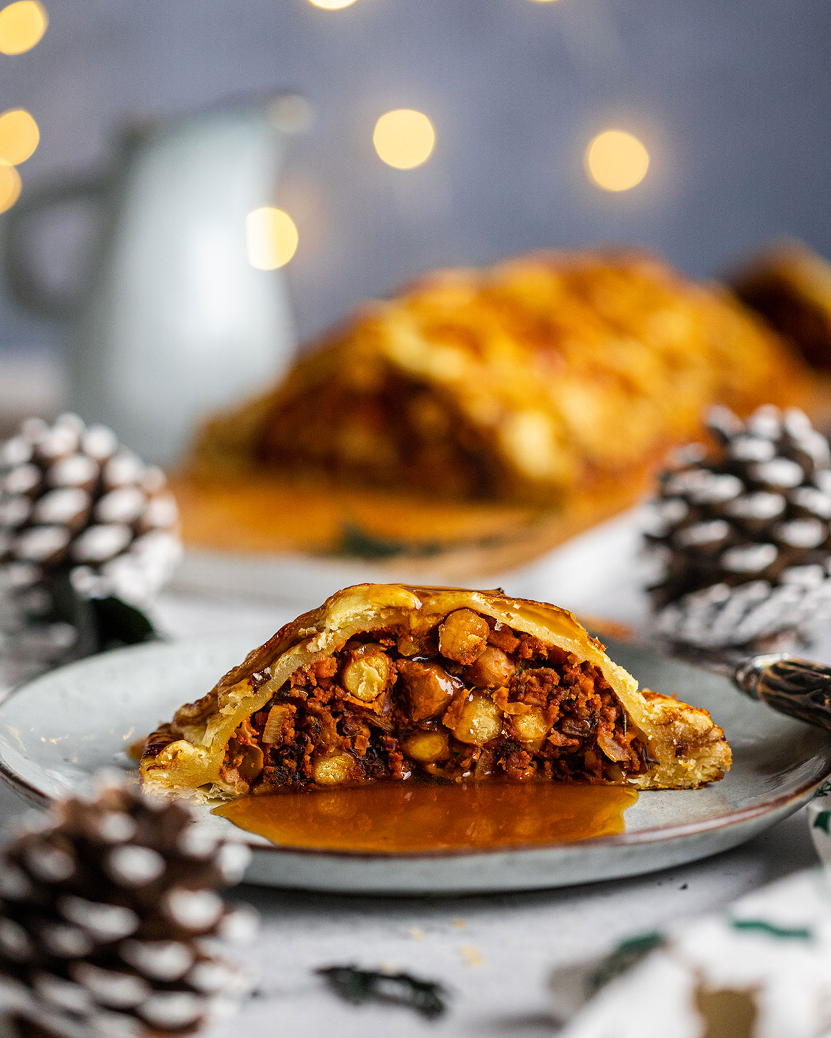 A slice of vegetable Wellington sitting on a light plate in front of the full Wellington decorated with pinecones and fairy lights.