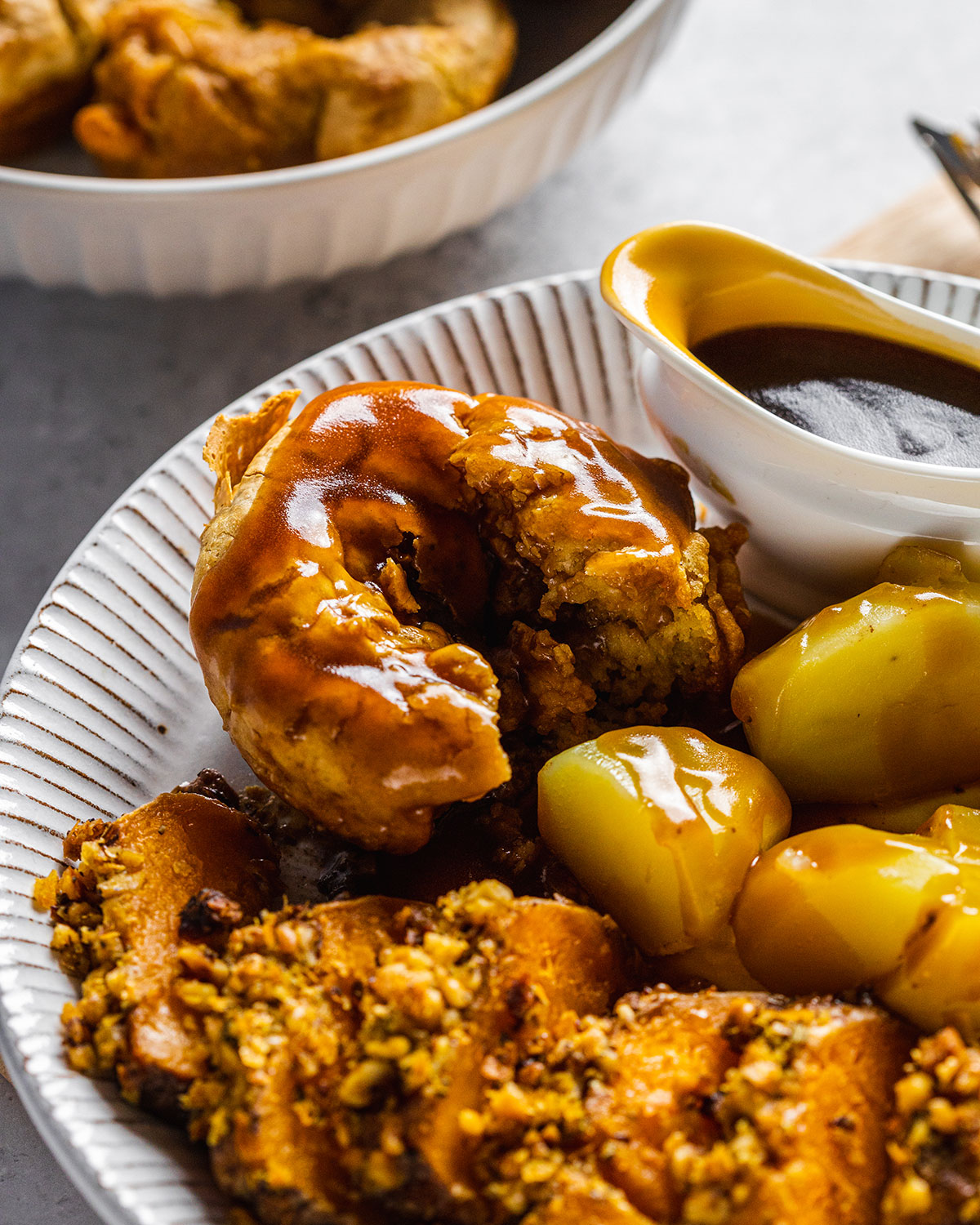 A vegan Yorkshire pudding on a dinner plate alongside potatoes and a vegetable roast, with a gravy boat on the side