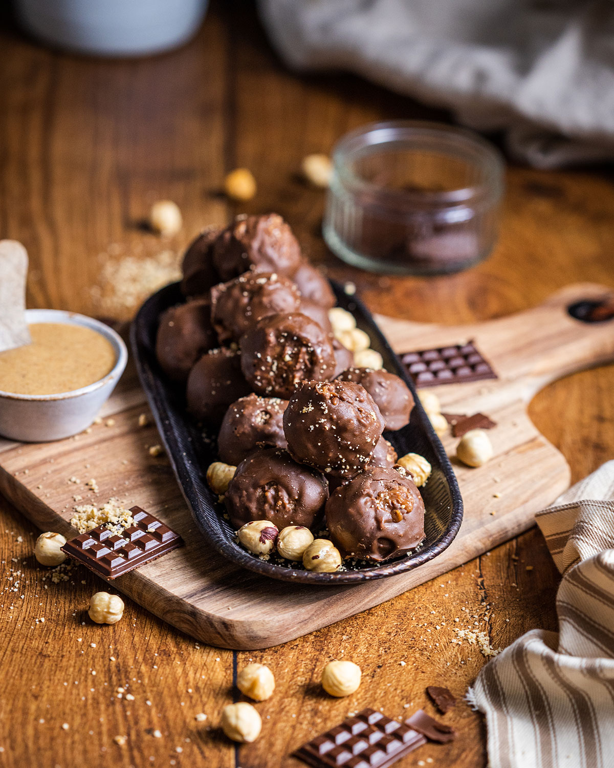 Small plate filled with vegan Ferrero Roché truffles sitting on a wooden chopping board surrounded by ingredients such as chocolate hazelnut butter and hazelnuts