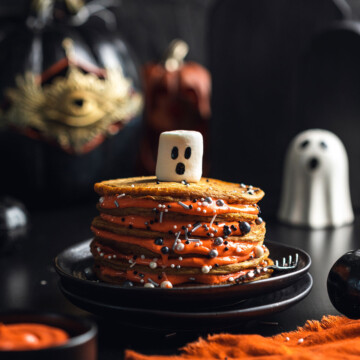 A stack of vegan pumpkin spice pancakes is shown on a black table with a marshmallow with ghost face on the top.