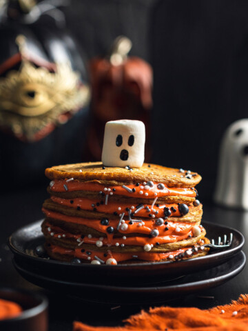 A stack of vegan pumpkin spice pancakes is shown on a black table with a marshmallow with ghost face on the top.