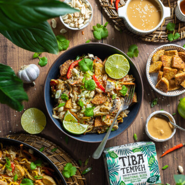 A portion of tempeh pad thai is served in a black bowl on a wooden table.