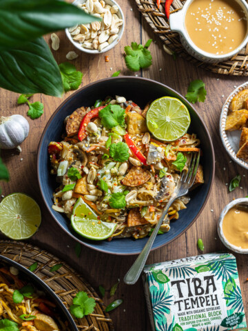 A portion of tempeh pad thai is served in a black bowl on a wooden table.