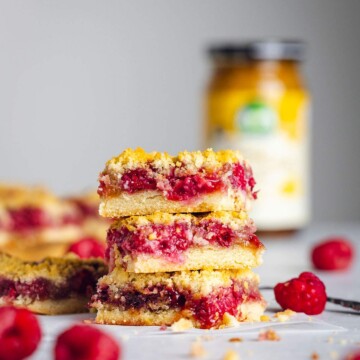 A stack of three raspberry crumble bars are sitting on a white table with their layers of crumble, raspberry, butterscotch and base clearly visible. More bars and a jar are visible in the background.