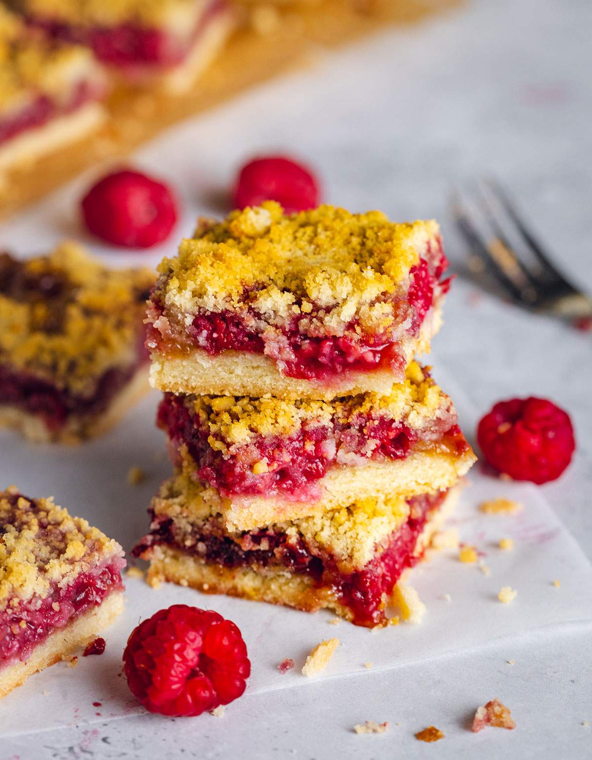 Three crumble bars filled with raspberries are stacked on a piece of white parchment on a table with more bars in the background.