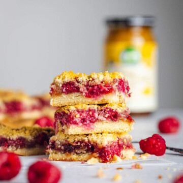 A stack of three raspberry crumble bars are sitting on a white table with their layers of crumble, raspberry, butterscotch and base clearly visible. More bars and a jar are visible in the background.