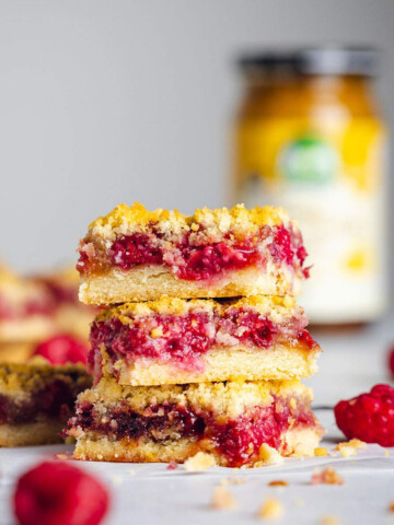 A stack of three raspberry crumble bars are sitting on a white table with their layers of crumble, raspberry, butterscotch and base clearly visible. More bars and a jar are visible in the background.