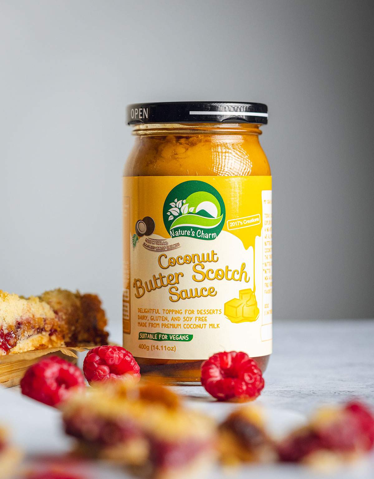 A jar of Nature's Charm Coconut Butterscotch Sauce is sitting on a table next to crumble bars and fresh raspberries.