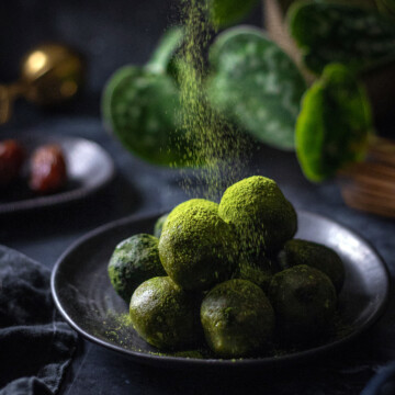 vegan matcha energy balls on a black plate with matcha powder being sprinkled onto them
