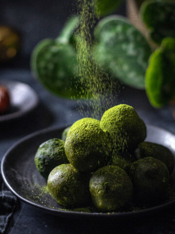 vegan matcha energy balls on a black plate with matcha powder being sprinkled onto them