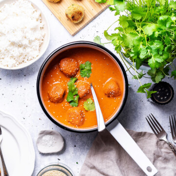 A white enamel saucepan filled with vegan falafel curry can be seen on a white table next to coriander, rice and other ingredients.