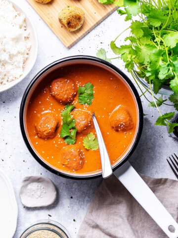 A white enamel saucepan filled with vegan falafel curry can be seen on a white table next to coriander, rice and other ingredients.