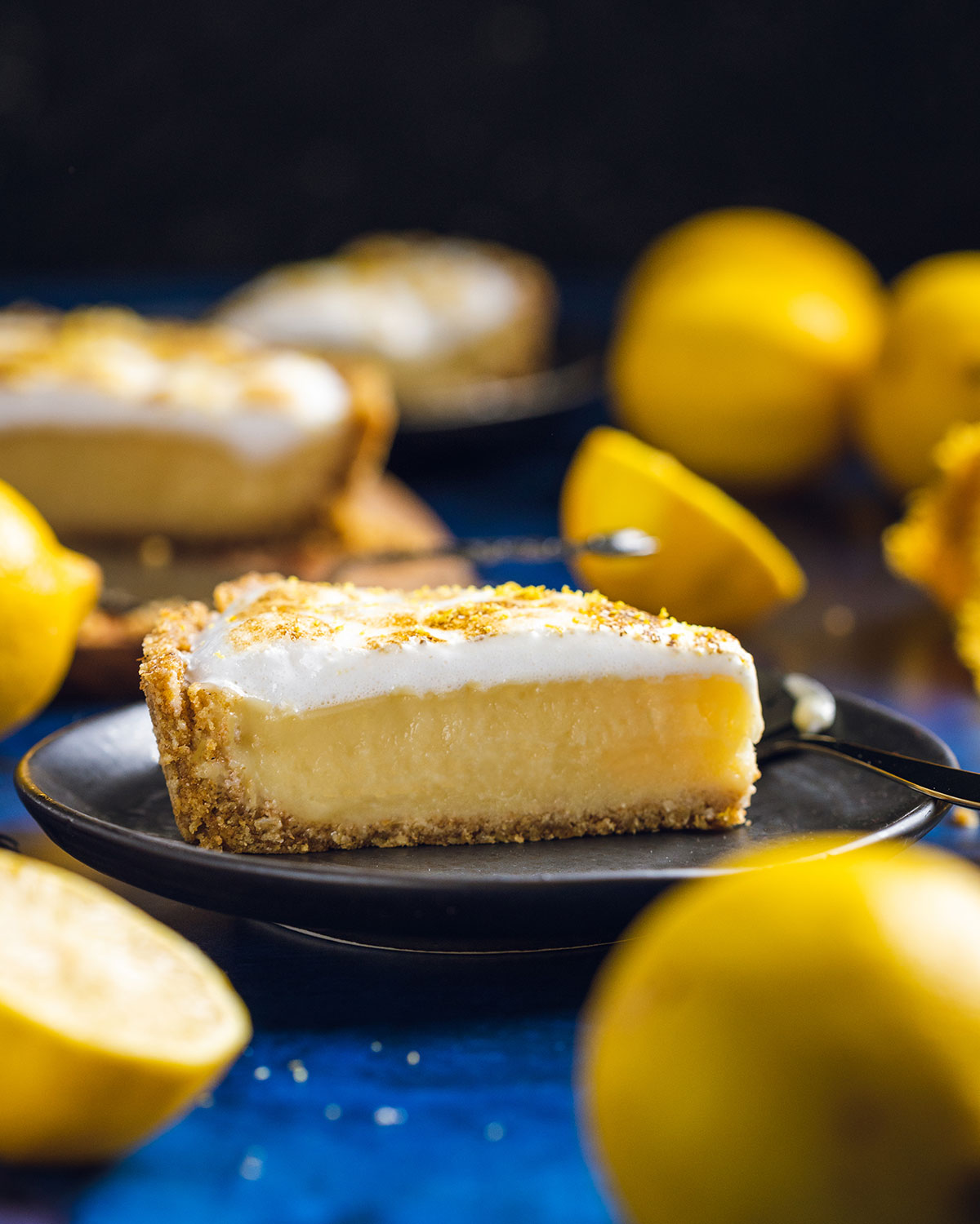 A slice of home-made vegan lemon meringue pie on a black plate surrounded by fresh lemons