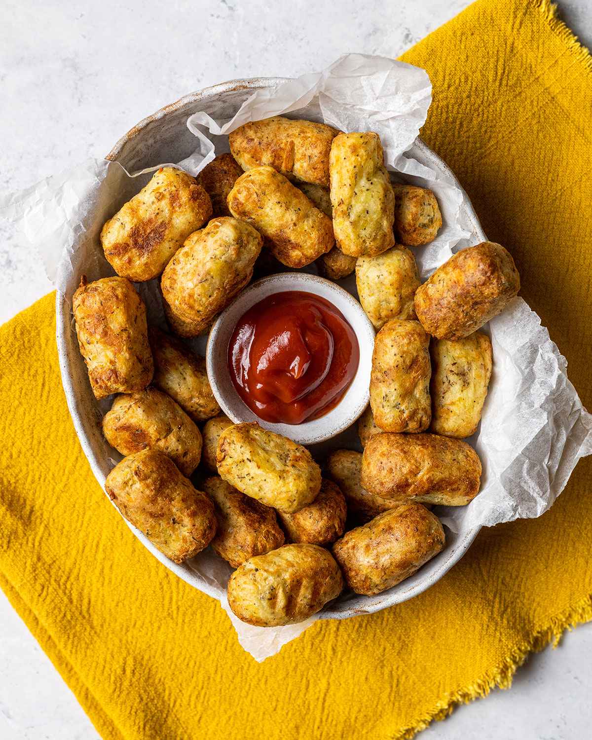 Air Fryer tater tots in a white bowl with ketchup in the middle, on a marble table with yellow napkin