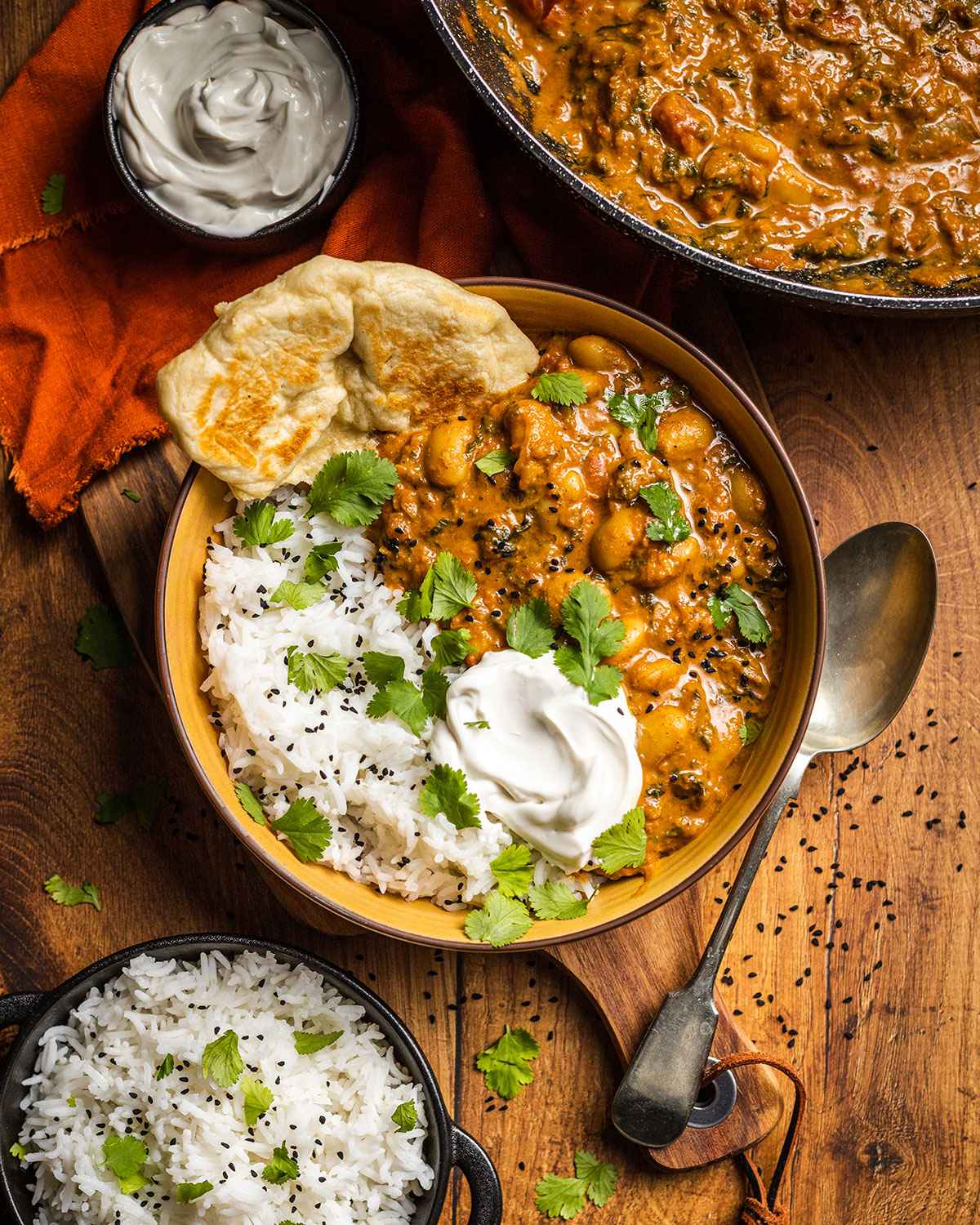 A yellow bowl is filled with rice and vegan butter bean curry on the dinner table. Next to the bowl there is a spoon, a dish with rice and a large pan with the curry. 