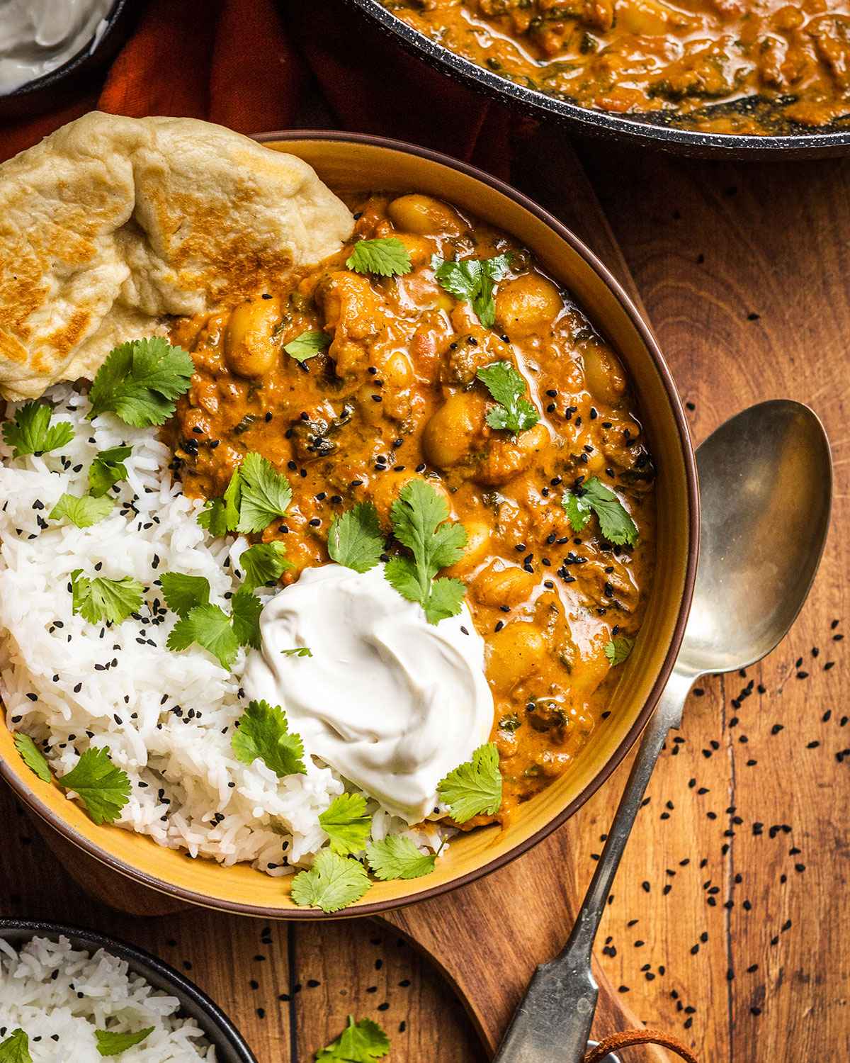 delicious vegan butter bean curry recipe in a yellow bowl alongside homemade naan bread, basmati rice, coconut yogurt and fresh coriander