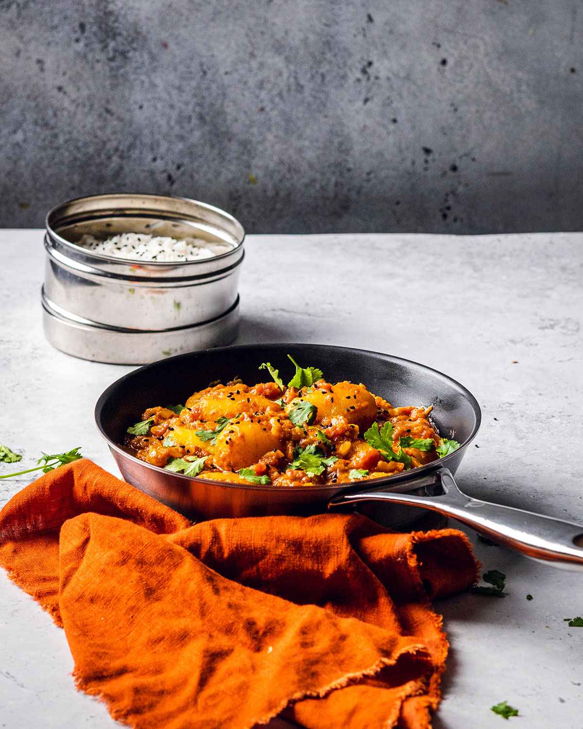 a skillet with potato curry and a pot of cooked rice on a white marble table with an orange napkin