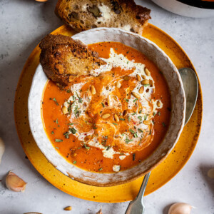 A bowl full of red pepper and sweet potato soup with a slice of toasted bread on the side.