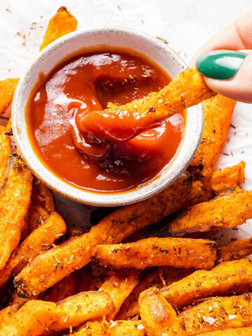 a hand dipping sweet potato fries into a small bowl of tomato ketchup