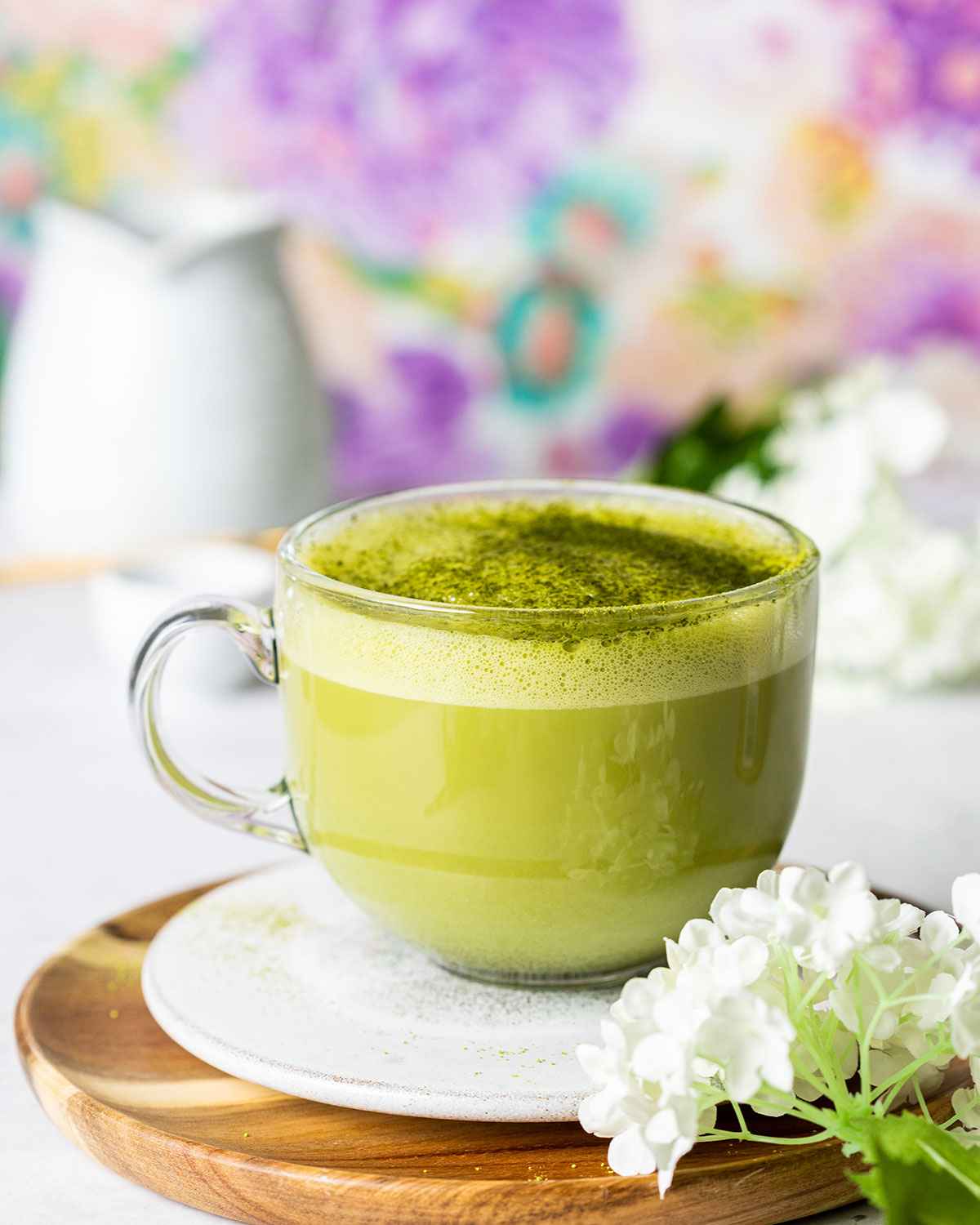 starbucks style matcha latte in a glass mug surrounded by white flowers and a milk jug blurry in the background