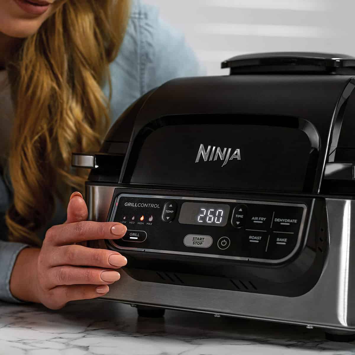 a ninja foodi 5-in-1 air fryer sitting on a kitchen counter, a woman with long hair leaning on the counter pressing a button on the device