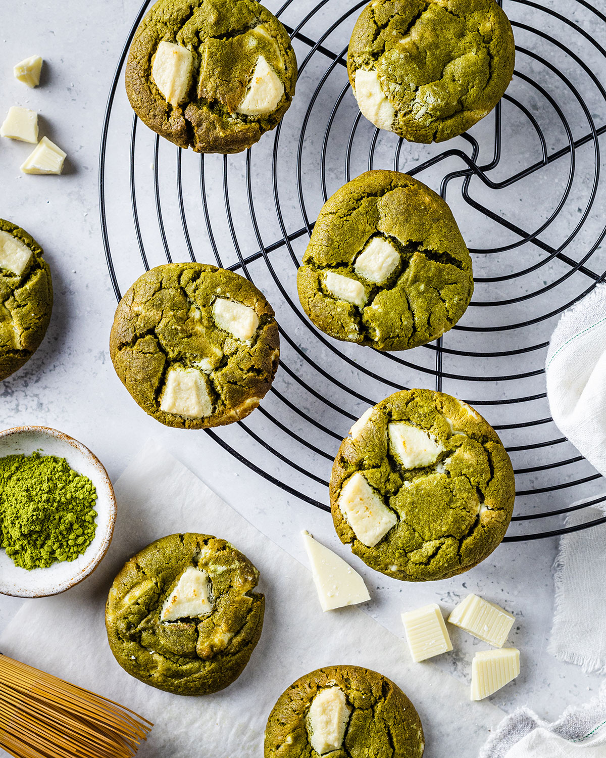 Baked Matcha green tea cookies it's white chocolate chips on a round cooling rack surrounded by ingredients such as white chocolate chips and green tea powder in a pinch pot