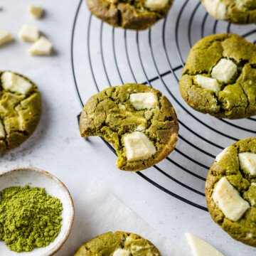 Matcha cookies on a cooling rack with a bite taken out of the cookie in the middle. Next to the cooling rack there is a pinch pot with green Matcha powder and a couple of pieces of white chocolate which is also used on the cookies.
