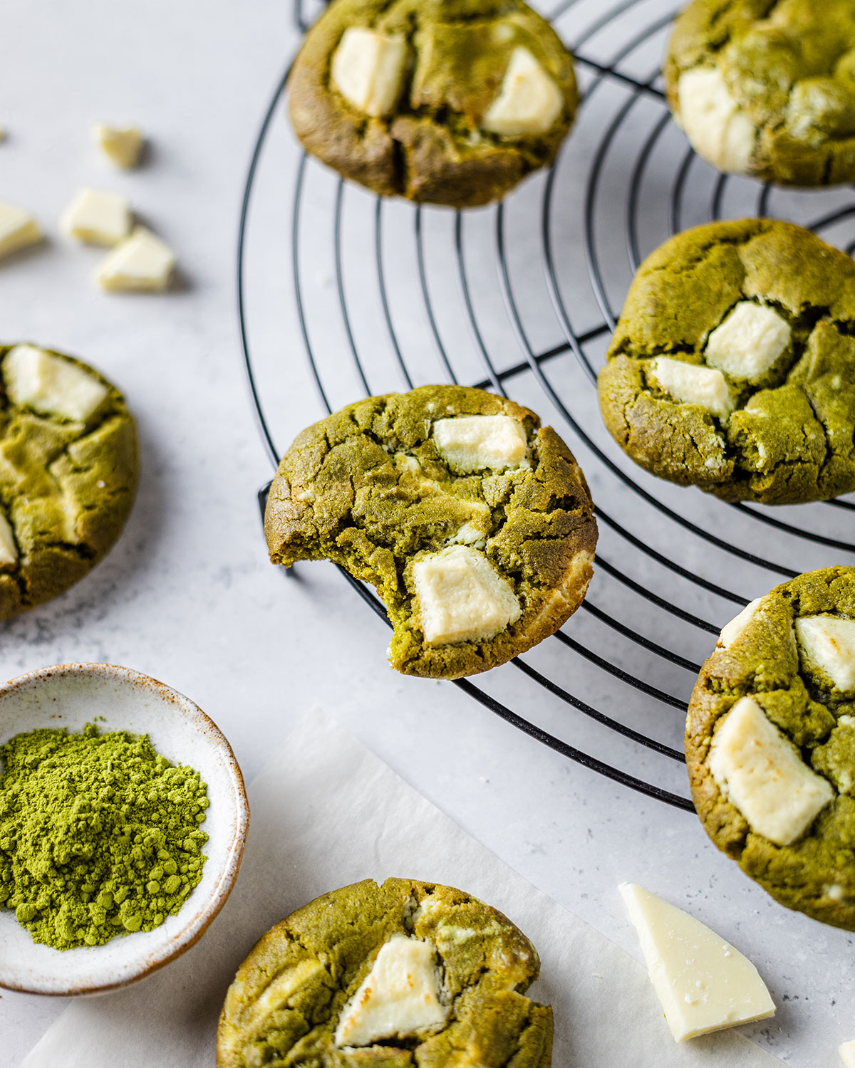 Matcha cookies on a cooling rack with a bite taken out of the cookie in the middle. Next to the cooling rack there is a pinch pot with green Matcha powder and a couple of pieces of white chocolate which is also used on the cookies.