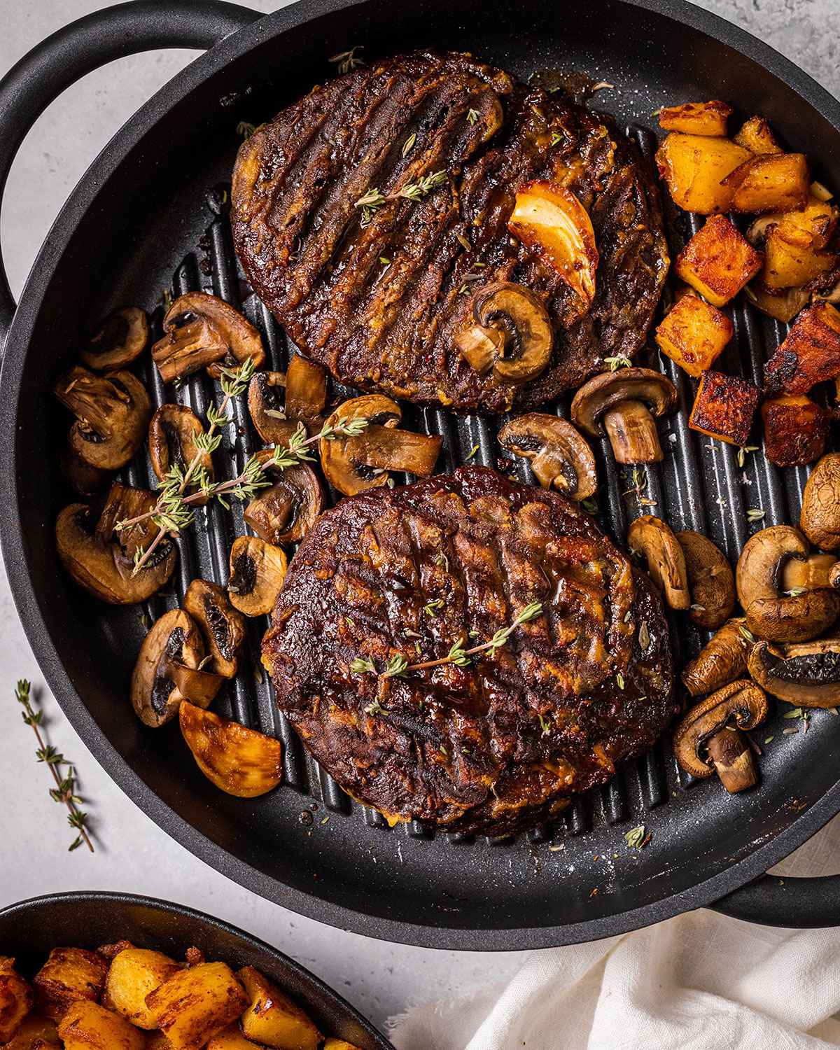 two vegan steaks in a skillet with mushrooms, thyme, garlic and roasted potato cubes