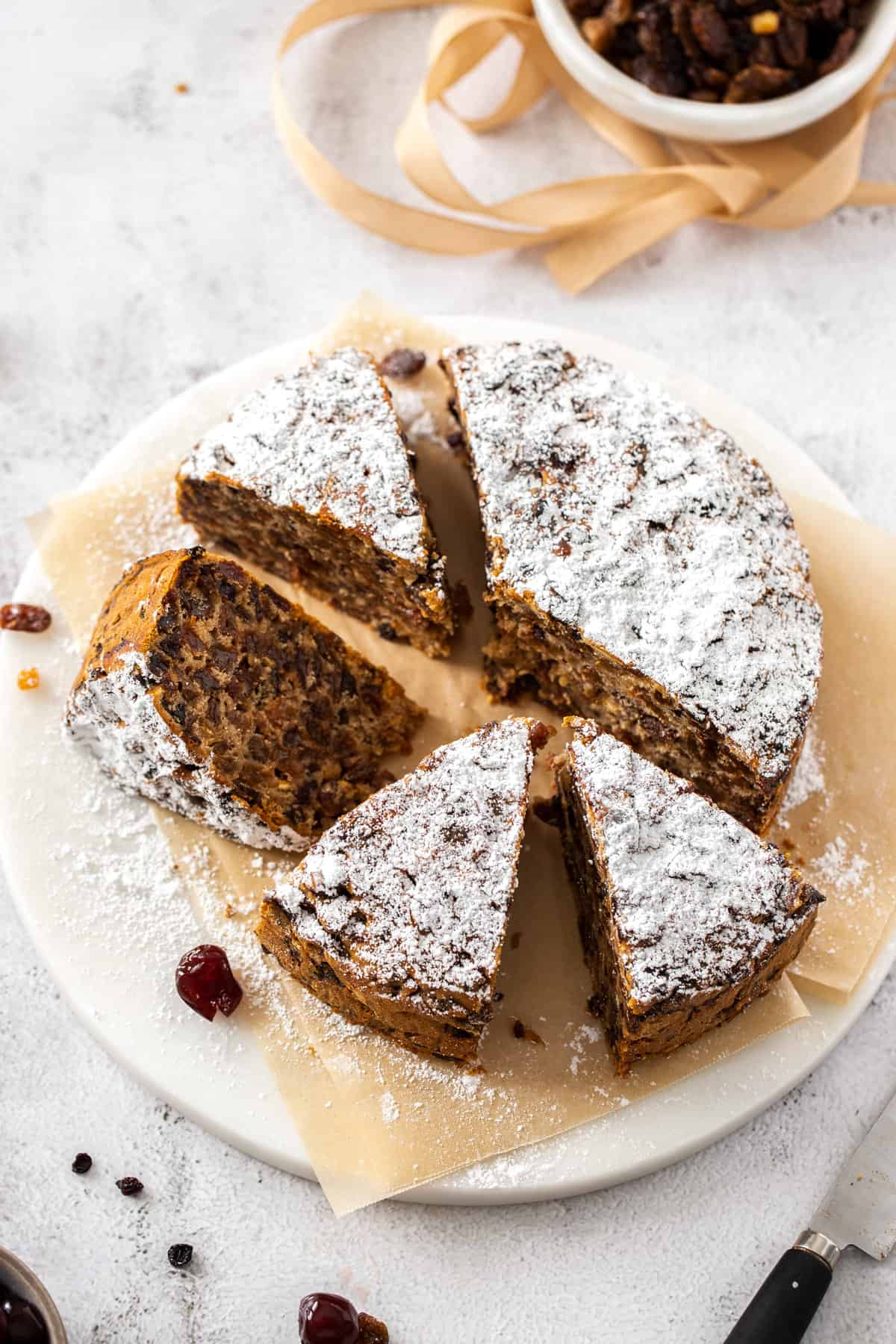 a round gluten free vegan fruitcake on a white platter with parchment paper. it's dusted with powdered sugar and 4 slices are cut out of it with one slice laying sideways next to the cake