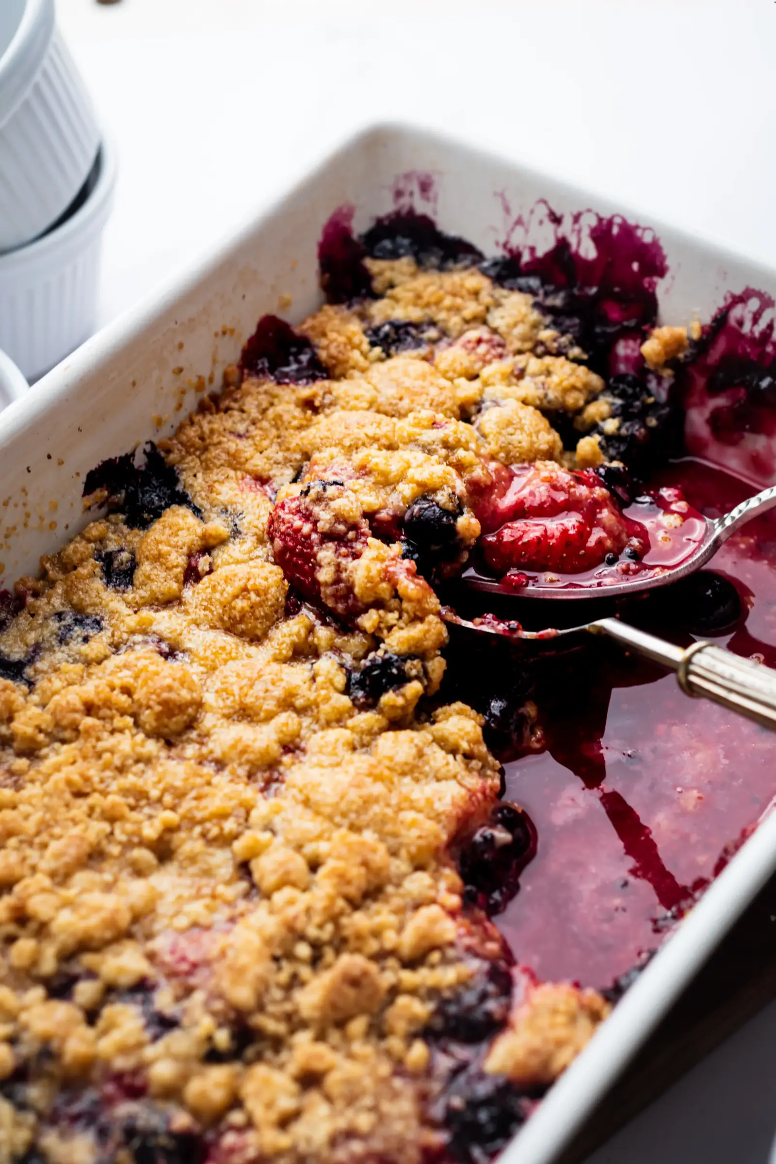 a large white rectangular oven dish with a vegan berry crumble, a spoon taking out the crumble