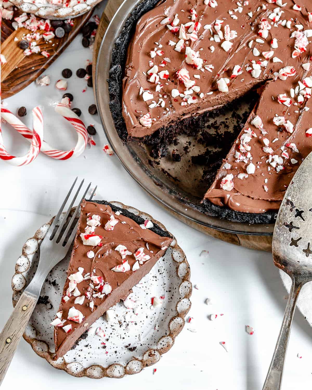 vegan chocolate peppermint pie made with silken tofu. The pie is served on a platter with a slice cut out of it, the slice it on a small white plate next to it and decorated with vegan candy cane pieces