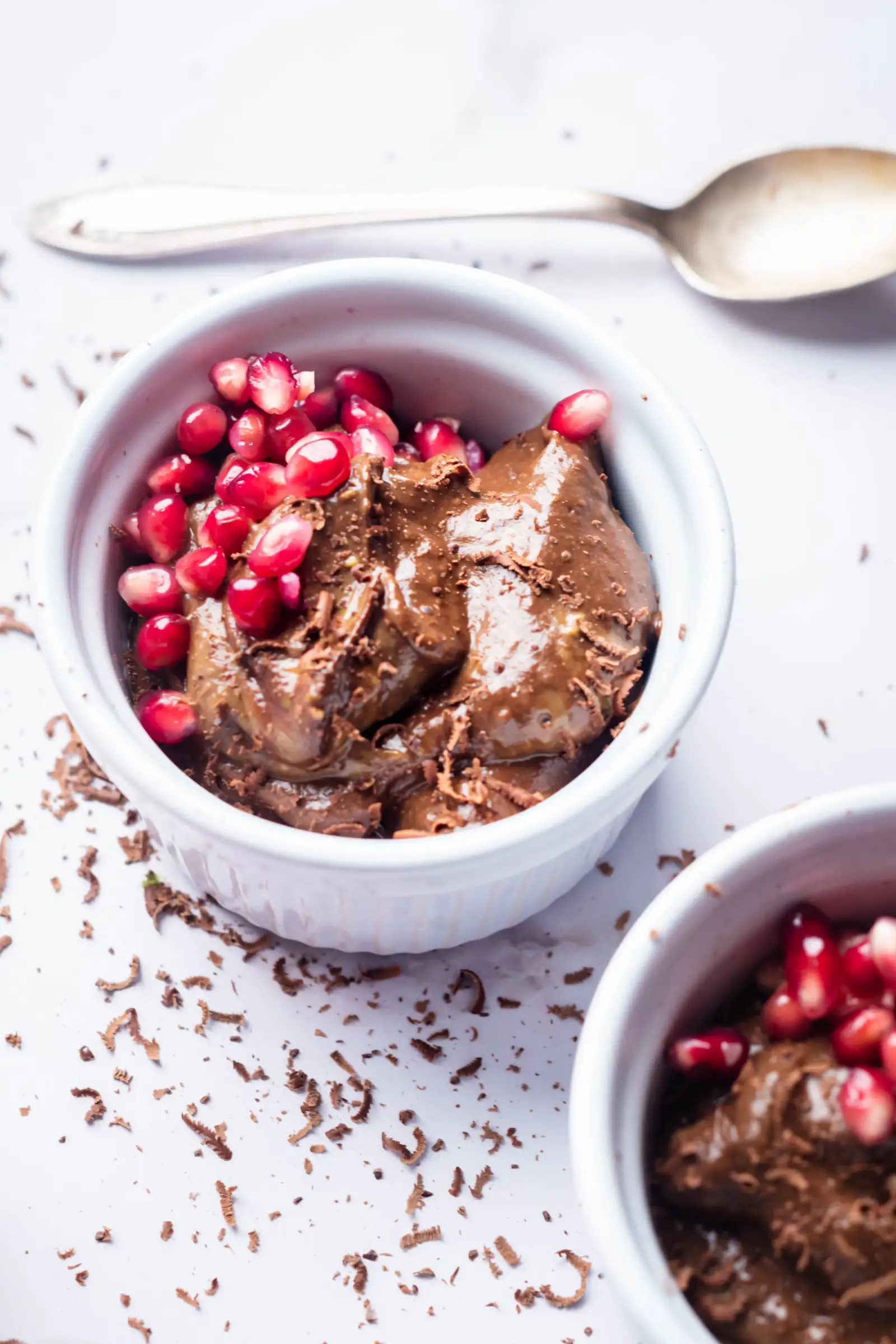 vegan chocolate avocado mousse in a white ramekin decorated with pomegranate seeds on top, another ramekin visible next to it and a spoon behind