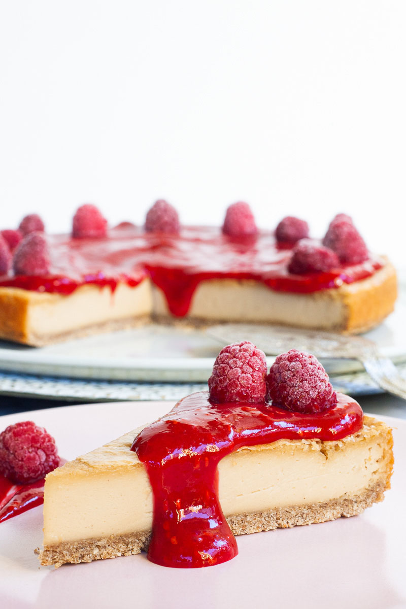 vegan baked cheesecake slice topped with raspberry sauce, a slice on a plate in the front with the rest of the cheesecake visible in the background