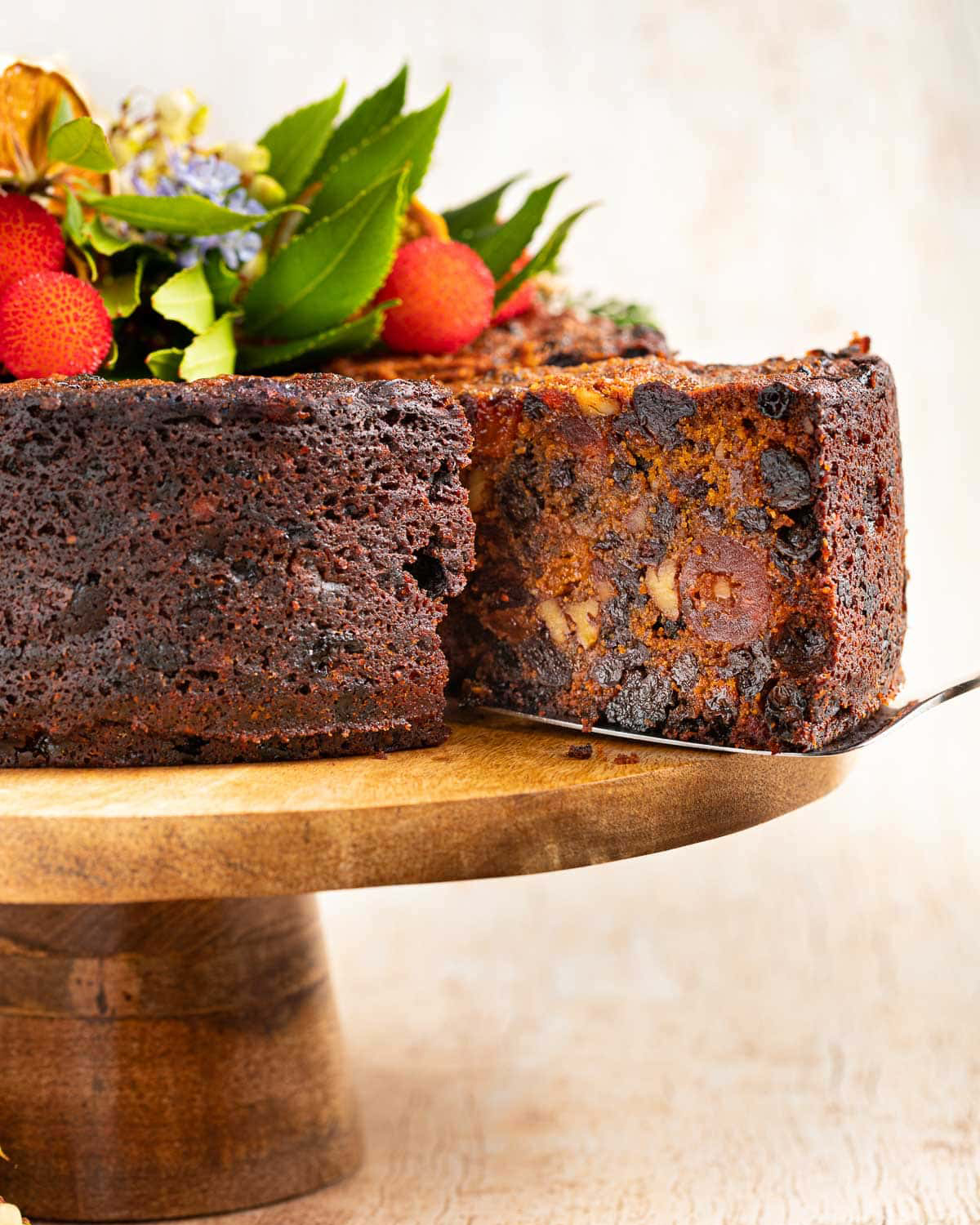 vegan christmas cake on a wooden cake tray, decorated with fruit on top and a cake lifter removing a slice from the cake, exposing the fruity and nutty interior