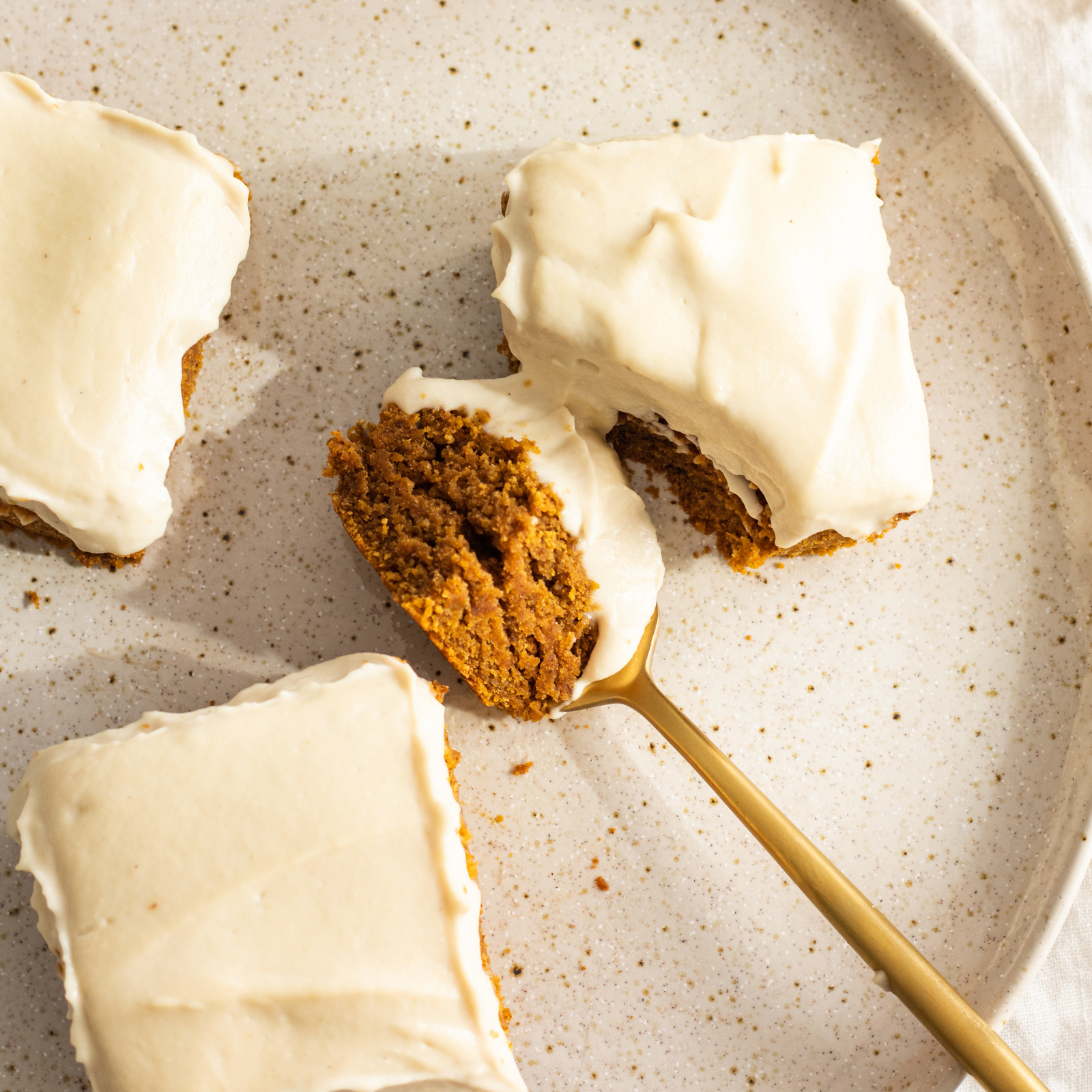 3 slices of vegan pumpkin cake topped with a vegan cream cheese frosting, seen from above on a white plate with a golden spoon taking a piece out of one of the slices.