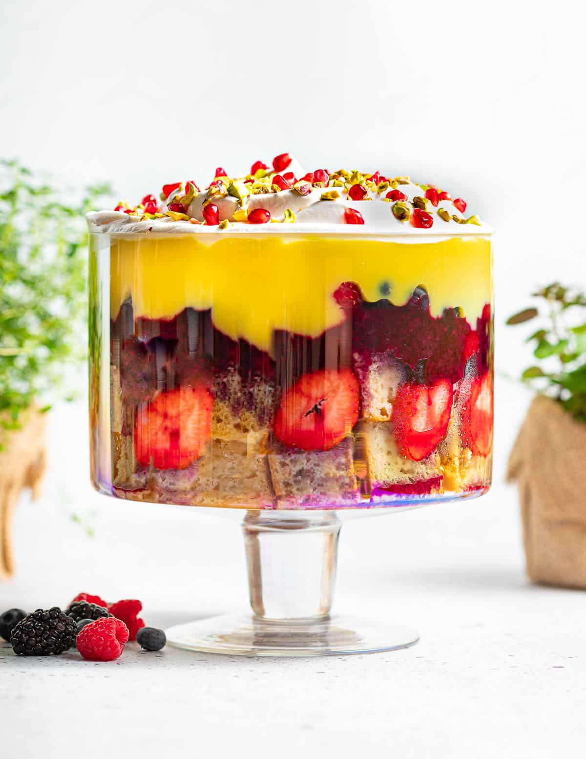 a vegan trifle served in a large glass dish for trifle. All the layers are clearly visible and you can spot strawberries and custard. Decorates with vegan whipped cream and pomegranate seeds on top.