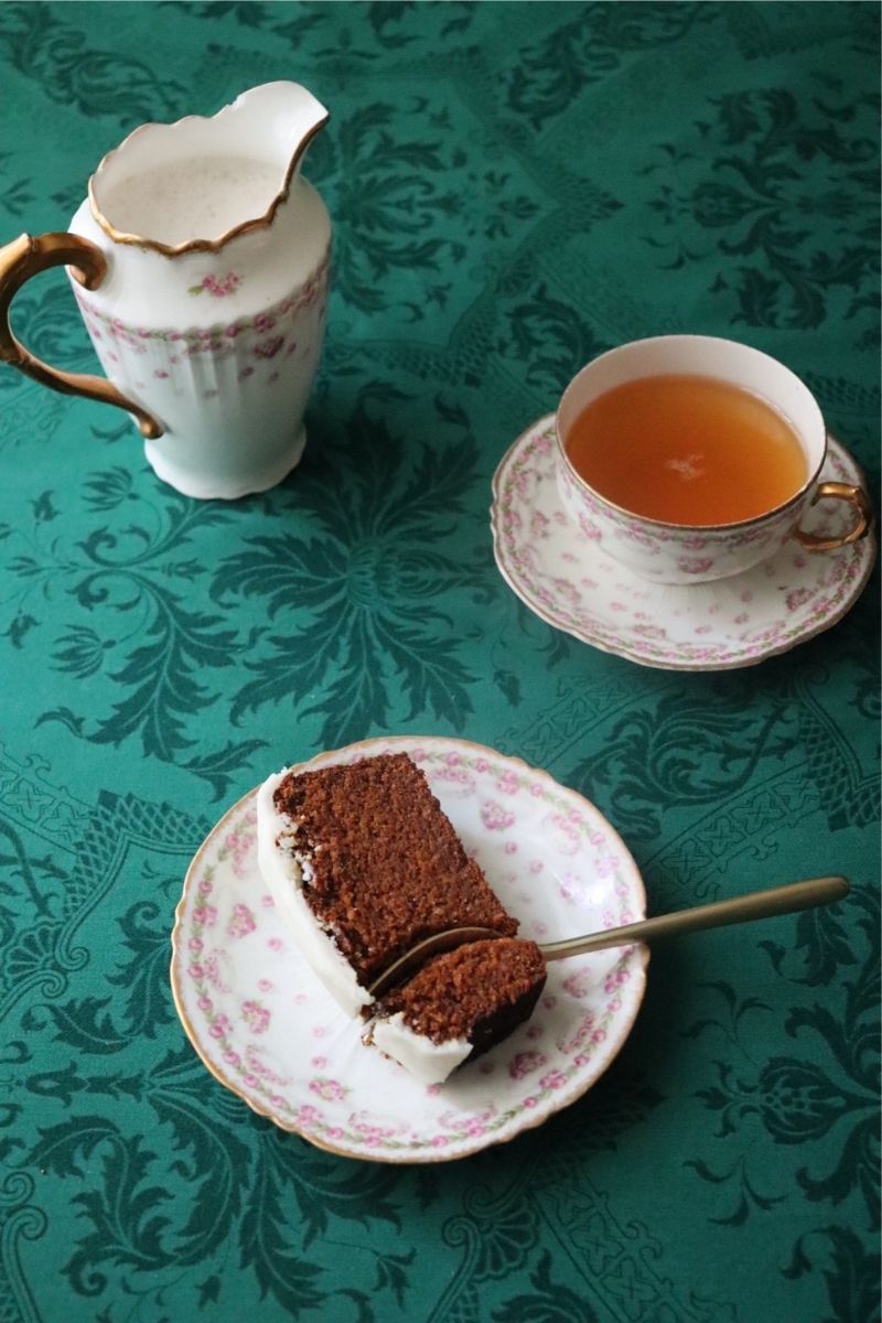 a slice of gluten free vegan gingerbread loaf on a dainty plate with a fork taking a piece of it. it's all sitting on a beautiful green tablecloth and a cup of tea is placed next to it